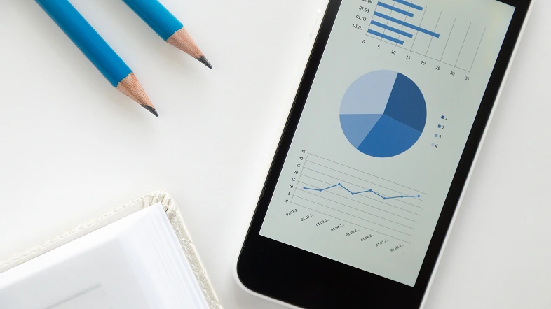 Смартфон с диаграммами, рядом карандаши, тетрадь на столе.