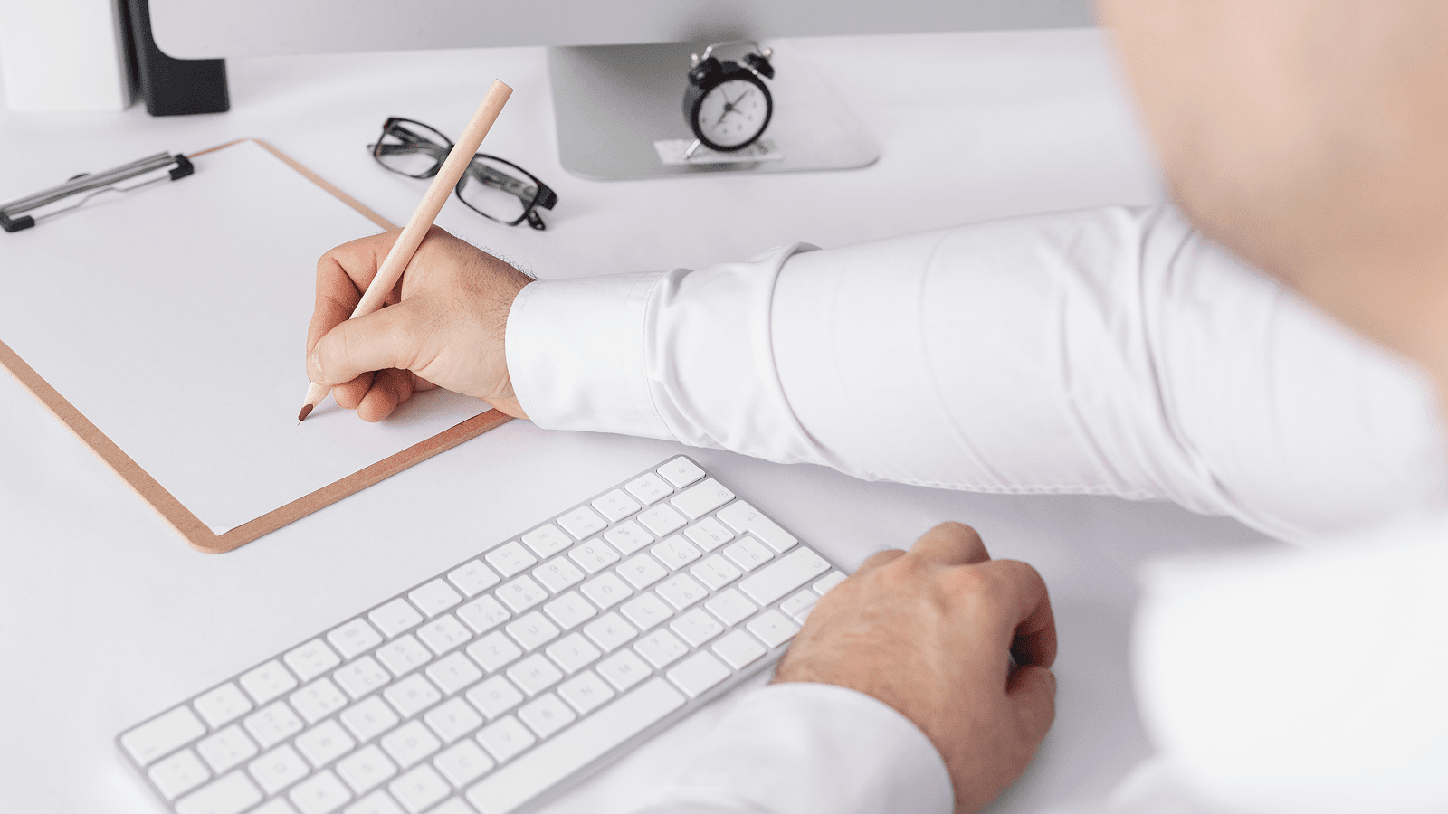 Человек пишет на клипборде рядом с клавиатурой и монитором