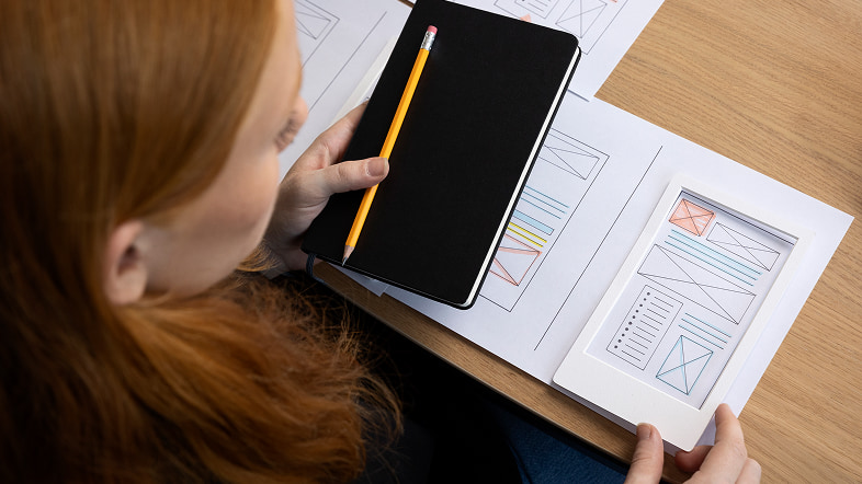 Женщина с карандашом держит смартфон над листами с набросками окон