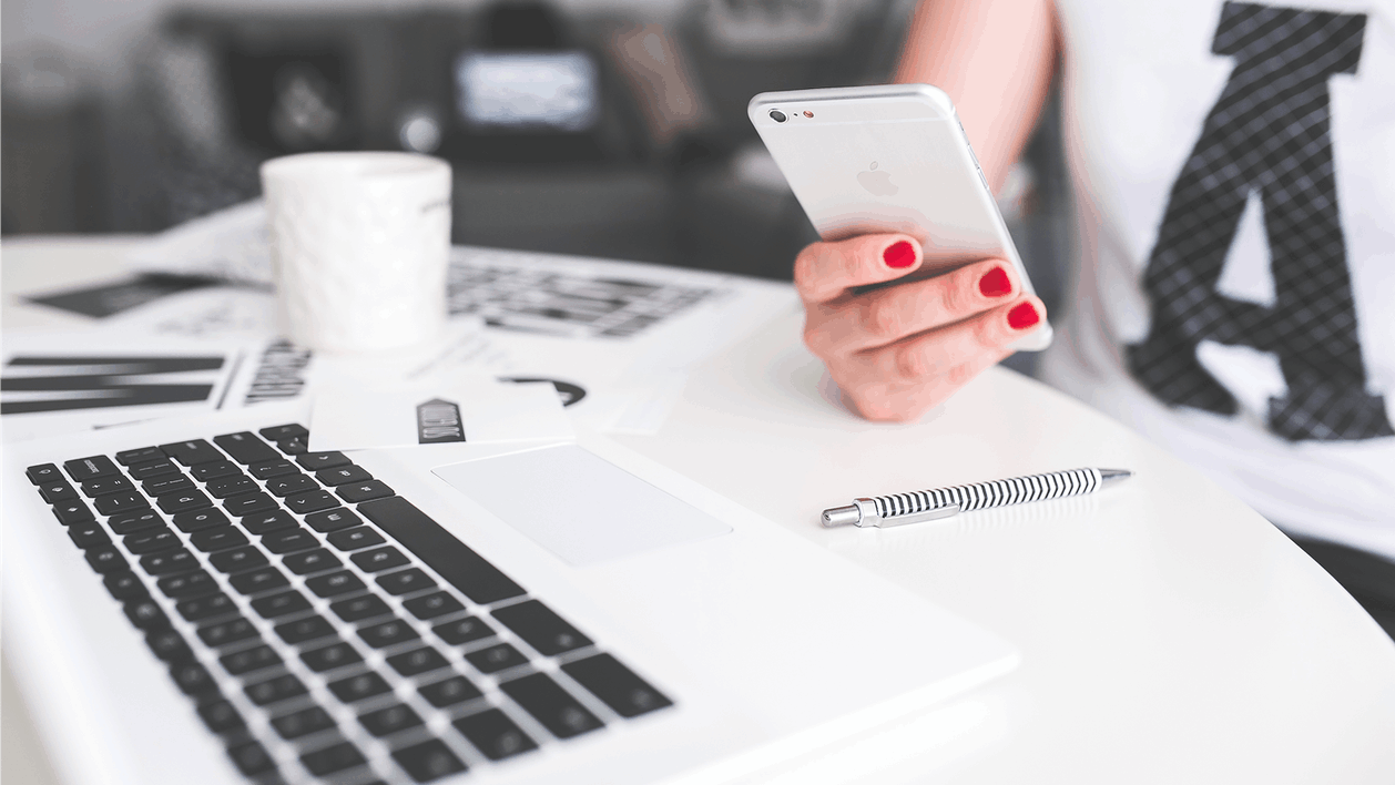 Женщина держит смартфон в руке за рабочим столом с ноутбуком и документами.