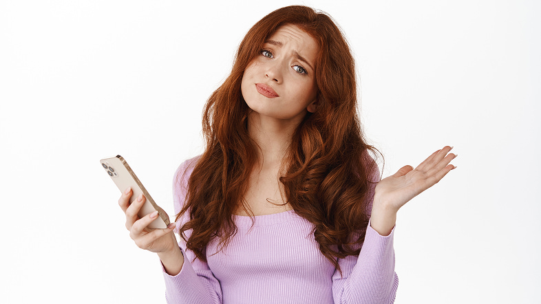 Молодая женщина с недоумением держит в руке смартфон